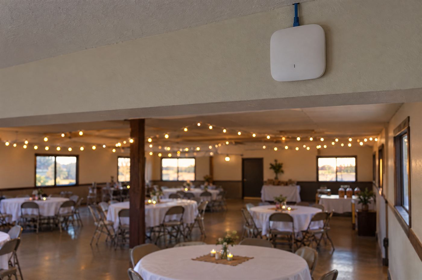 An empty event hall with round tables and string lights, and a Wi-Fi access point mounted high on the wall.